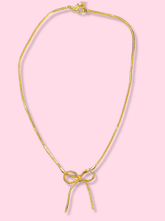 Gold chain necklace with a delicate bow pendant against a pink background.