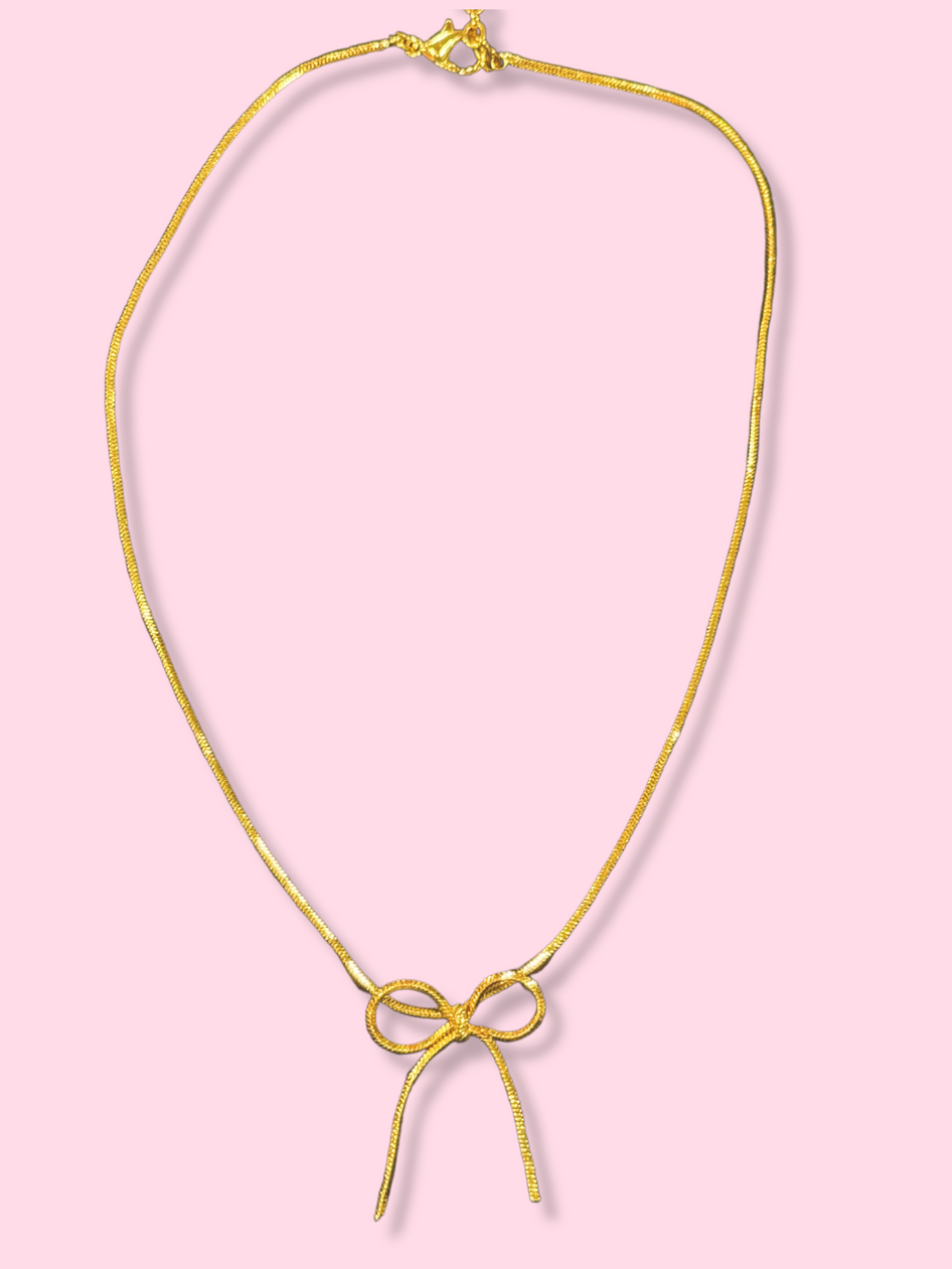 Gold chain necklace with a delicate bow pendant against a pink background.
