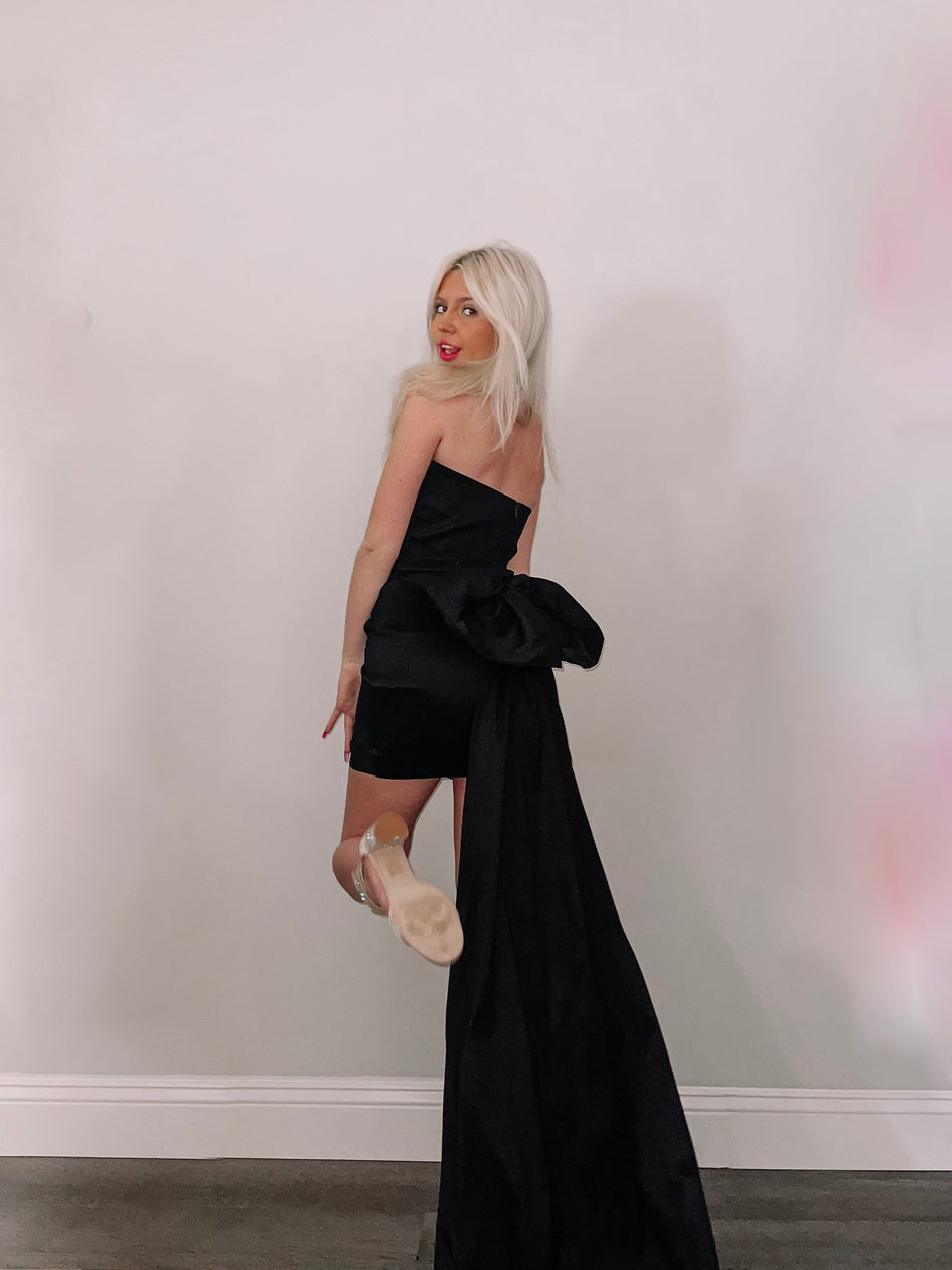 Black mini dress with dramatic train, modeled against a white wall.
