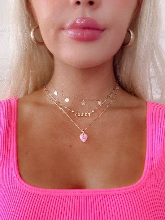 Kali Heart Necklace: Pink enamel heart pendant with gold chain, layered with delicate gold necklaces.
