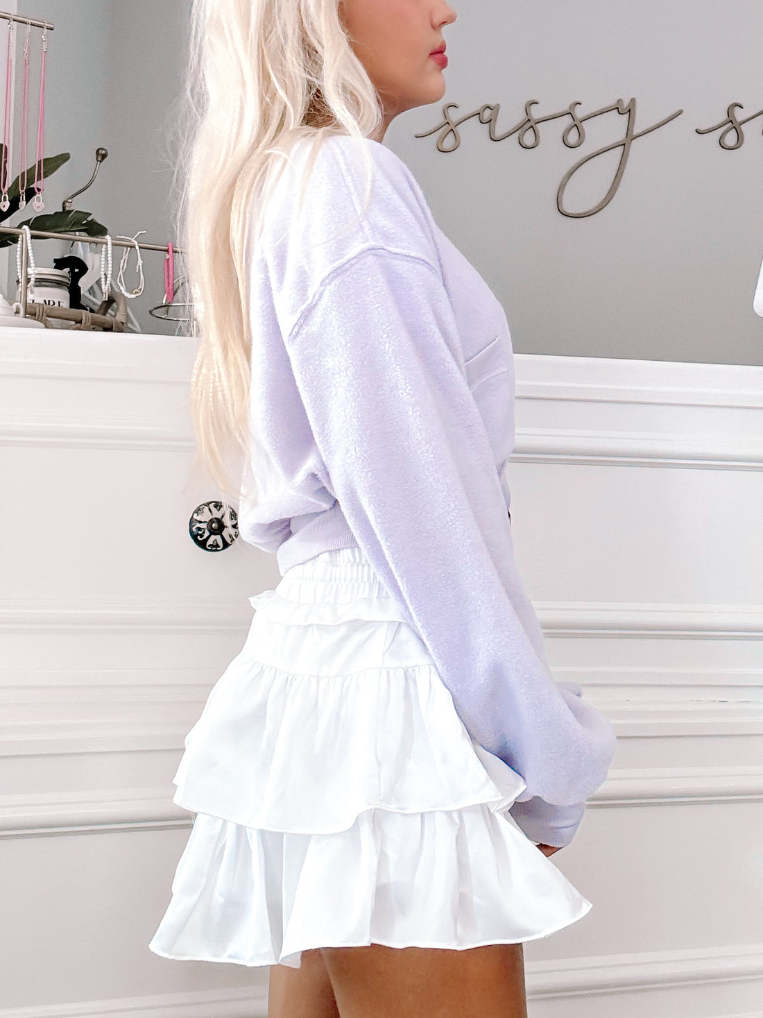 White tiered ruffle mini skirt paired with a lavender sweater against a white paneled wall.
