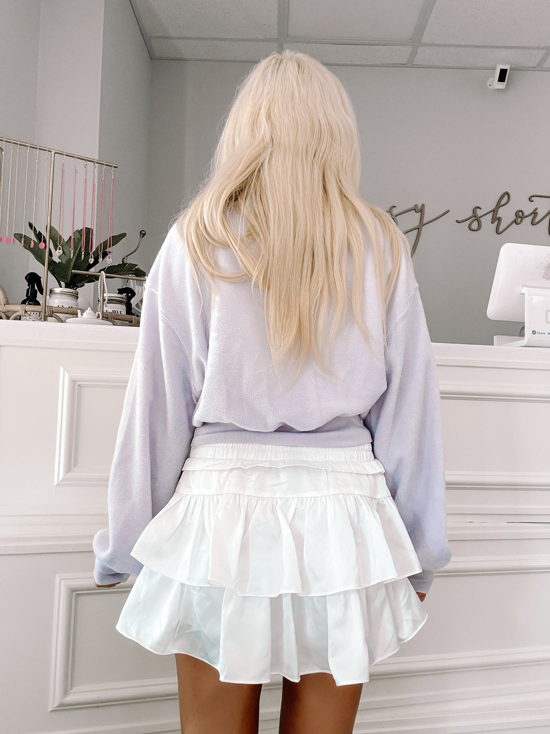 Lilac sweater and white tiered mini skirt shown from behind in a retail setting.
