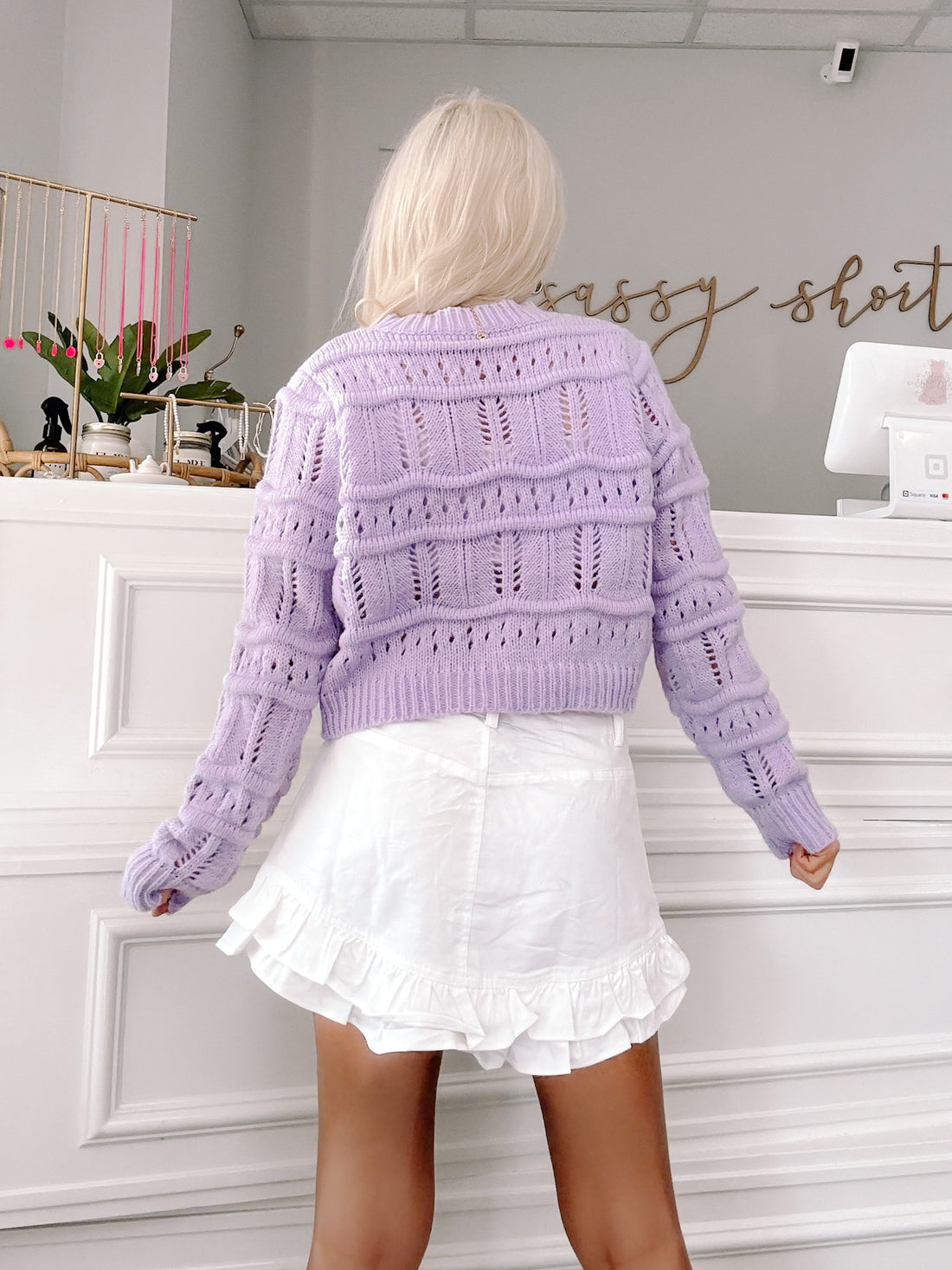 Lilac open-knit cropped cardigan, styled with a white ruffle skirt.
