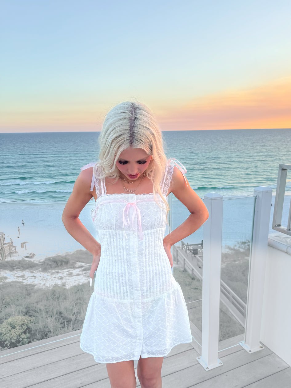 White eyelet mini dress with pink ribbon accents, worn on a beachside balcony at sunset.
