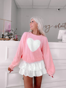 Pink knit sweater featuring a large white heart, modeled in a boutique setting.
