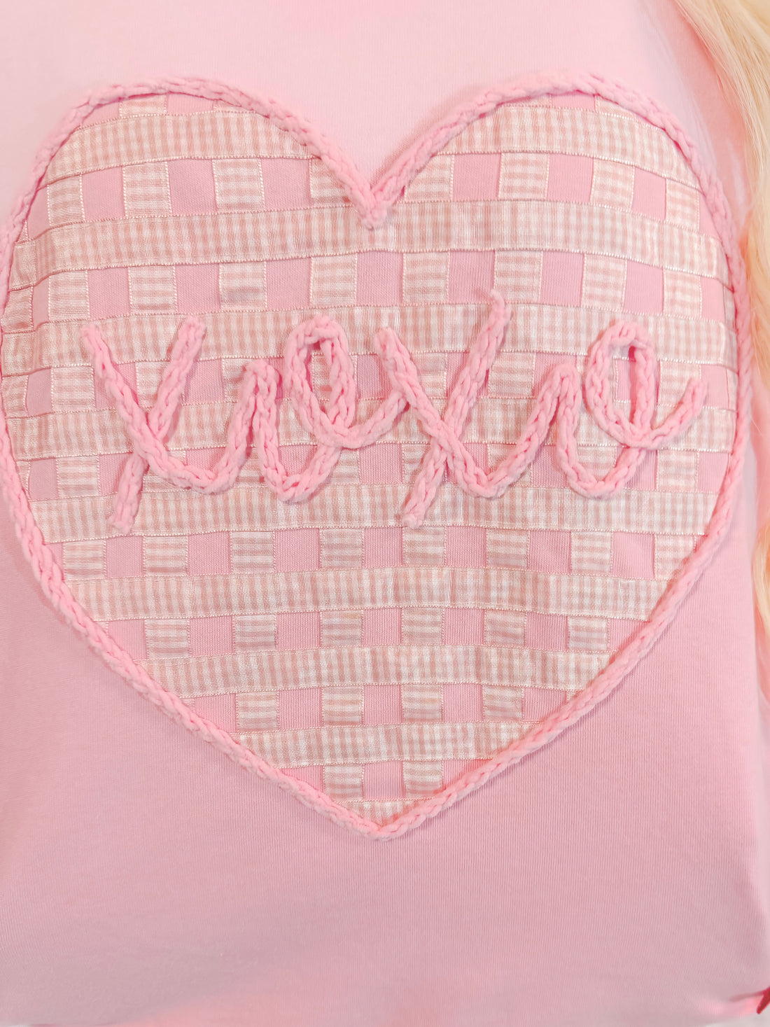 Pink crewneck sweater detail: heart-shaped design with woven ribbon and yarn "love" lettering.

