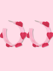 Pink hoop earrings adorned with crimson hearts, shown against a light pink background.
