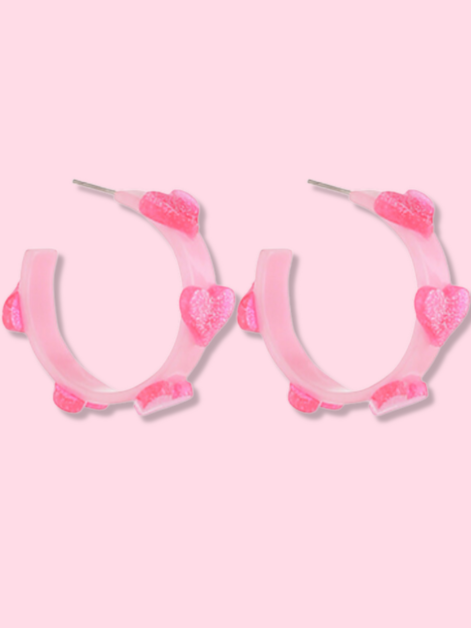 Pink hoop earrings adorned with small, sparkly hearts against a light pink background.
