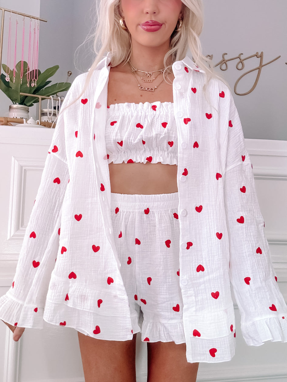White three-piece pajama set with small red hearts, featuring a button-down shirt and ruffled shorts.
