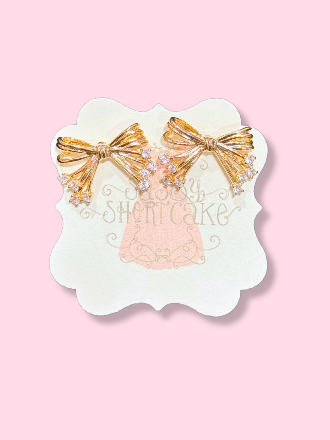 Gold Lucille Bow earrings with sparkling crystals, displayed on a pink background.
