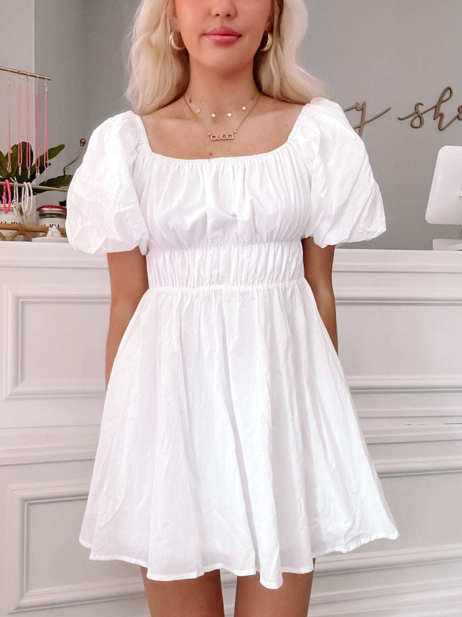 White mini dress with puff sleeves and a fitted waist, shown on a model in a bright interior.
