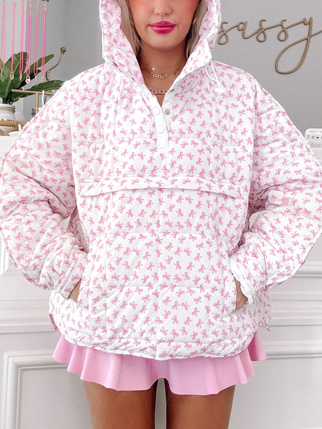 White quilted pullover with pink bow print, featuring a hood and pockets.
