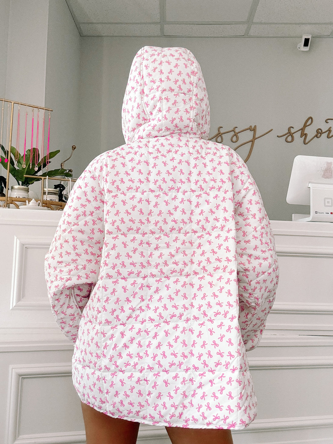 White hooded puffer jacket with a pink bow print, shown from the back in a shop setting.
