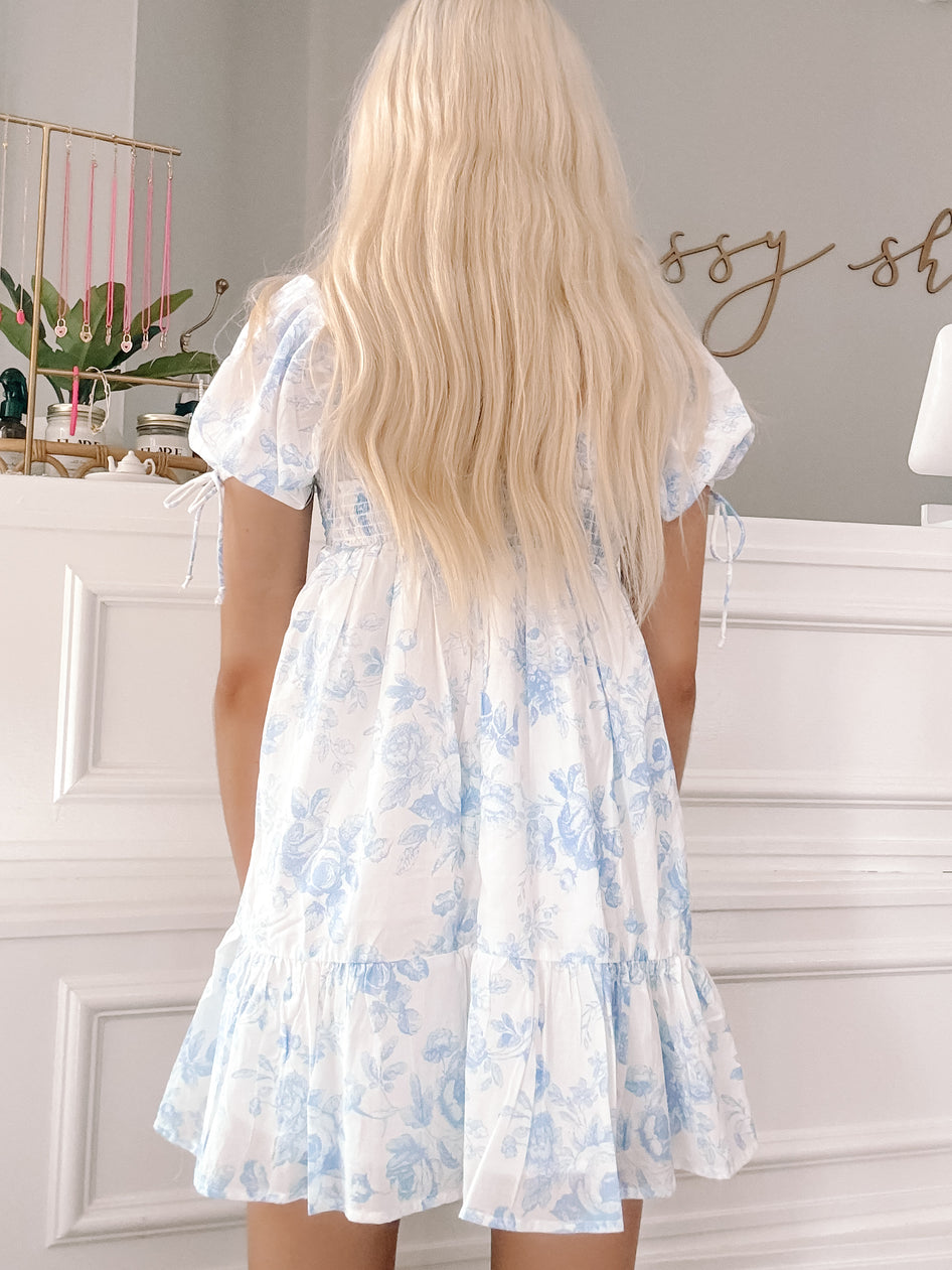 Blue floral mini dress with puff sleeves and tiered ruffle hem, shown from the back against a white wall.
