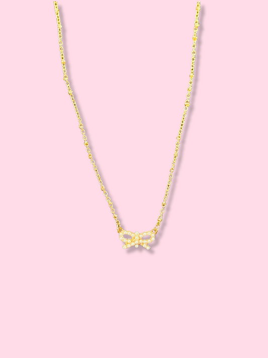 Dainty gold chain necklace with a small pearl bow pendant against a pink background.