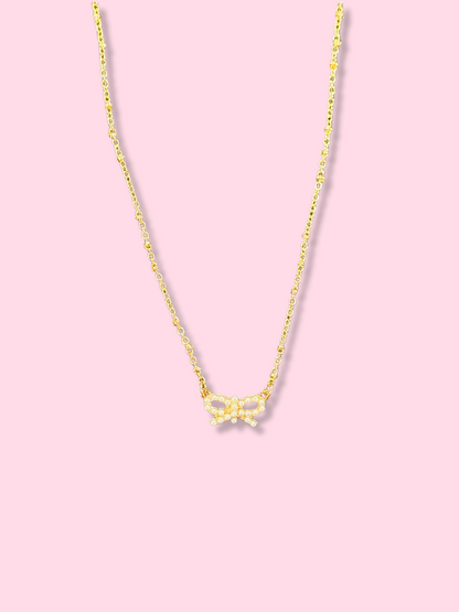 Dainty gold chain necklace with a small pearl bow pendant against a pink background.
