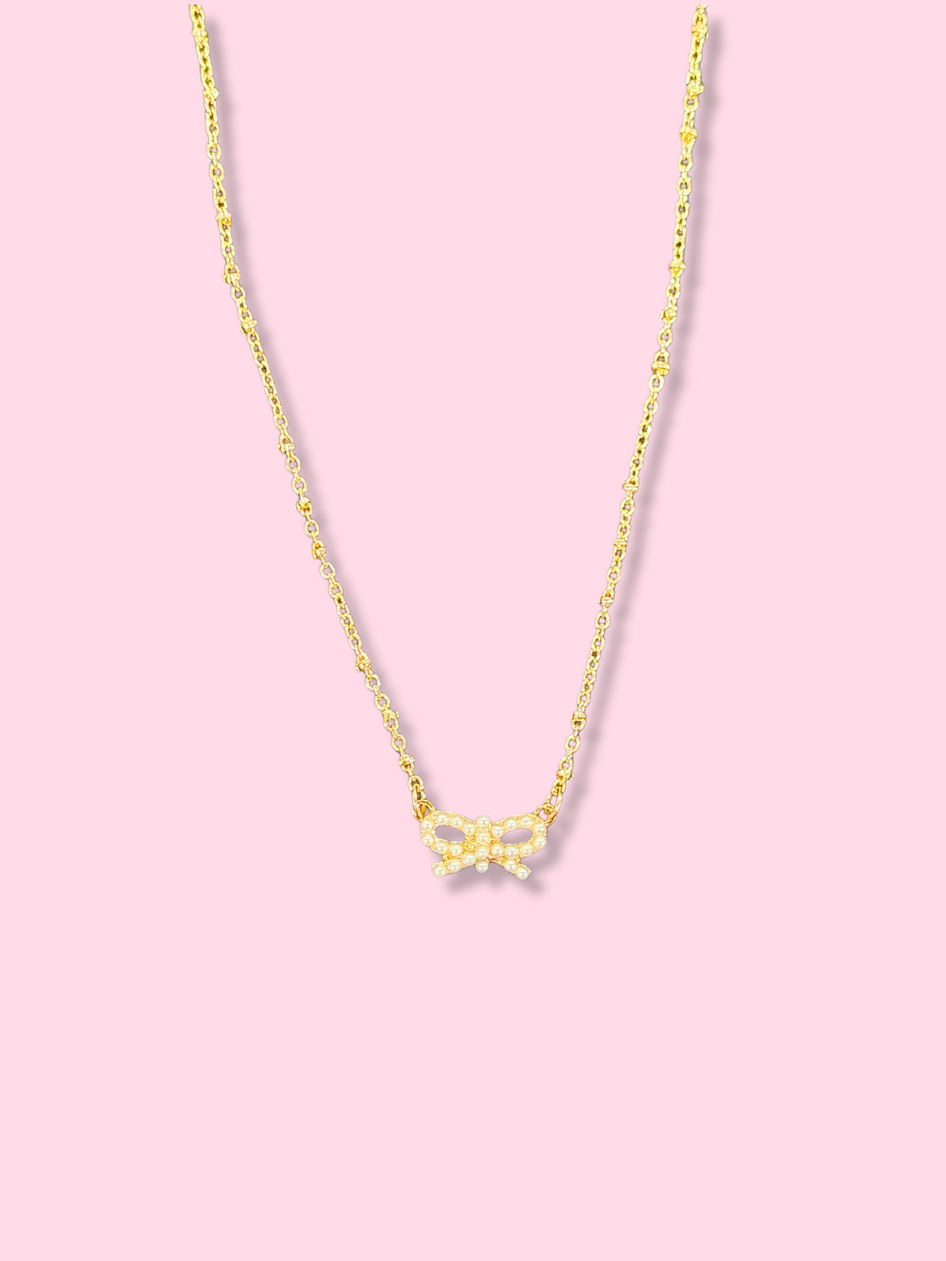 Dainty gold chain necklace with a small pearl bow pendant against a pink background.
