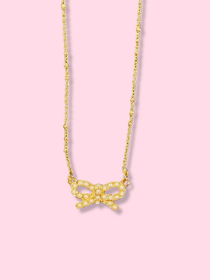 Gold chain necklace with delicate pearl bow pendant against a pink backdrop.
