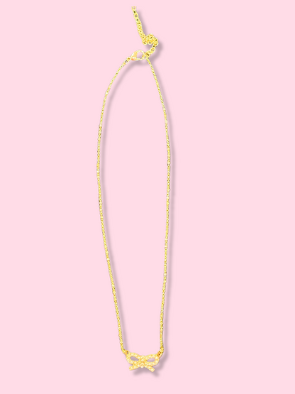 Gold chain necklace with a small pearl bow pendant against a pink background.
