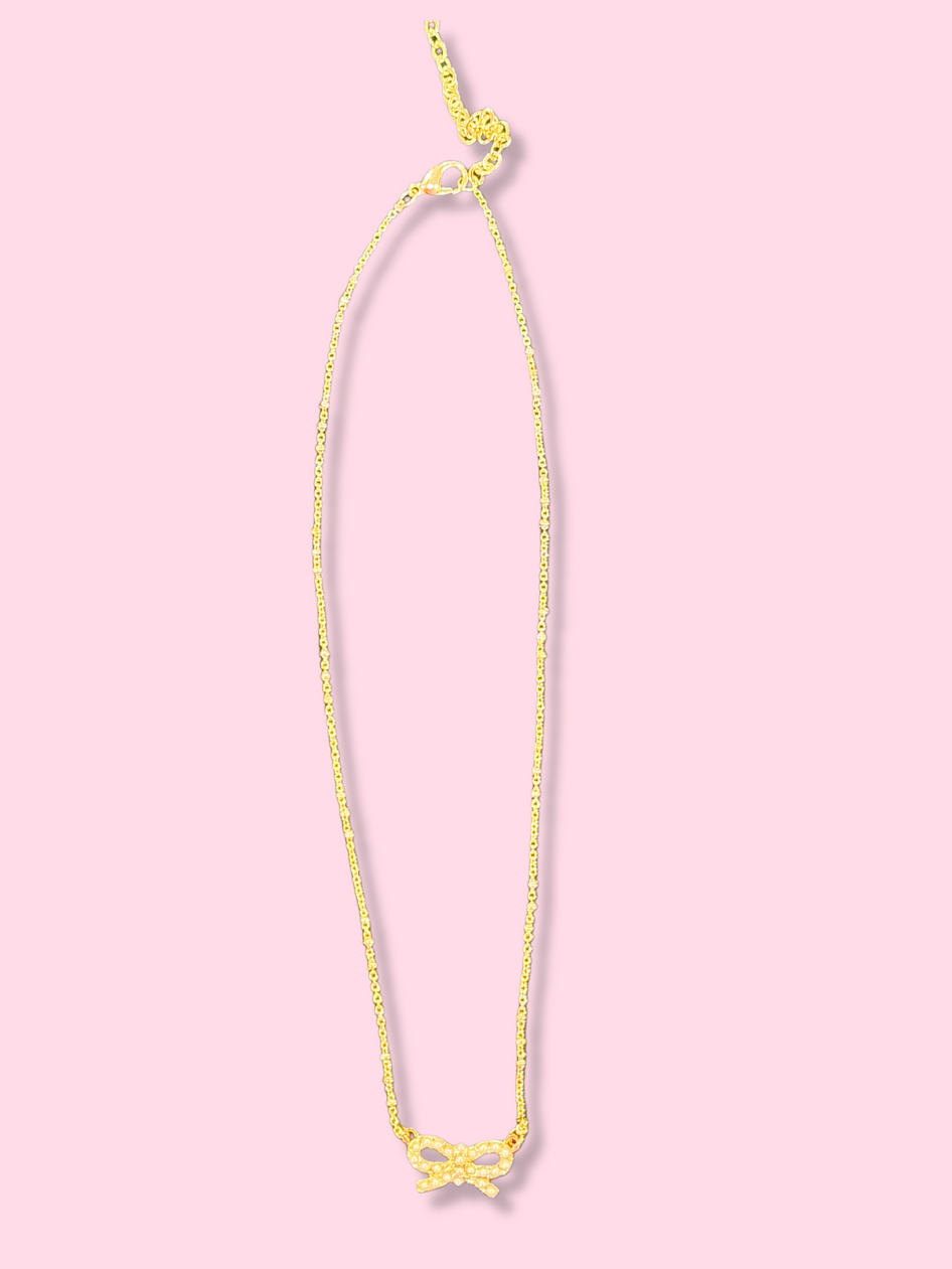 Gold chain necklace with a small pearl bow pendant against a pink background.
