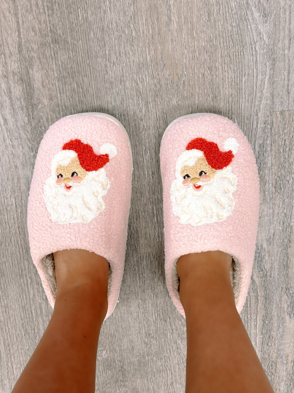Pink fuzzy slippers featuring an embroidered Santa Claus design, shown on feet against a wood floor.
