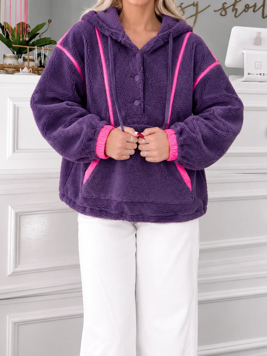 Purple fuzzy sherpa pullover with pink contrast piping, hood, button neckline and kangaroo pocket styled with white pants. Plum Pretty Pullover from Sassy Shortcake