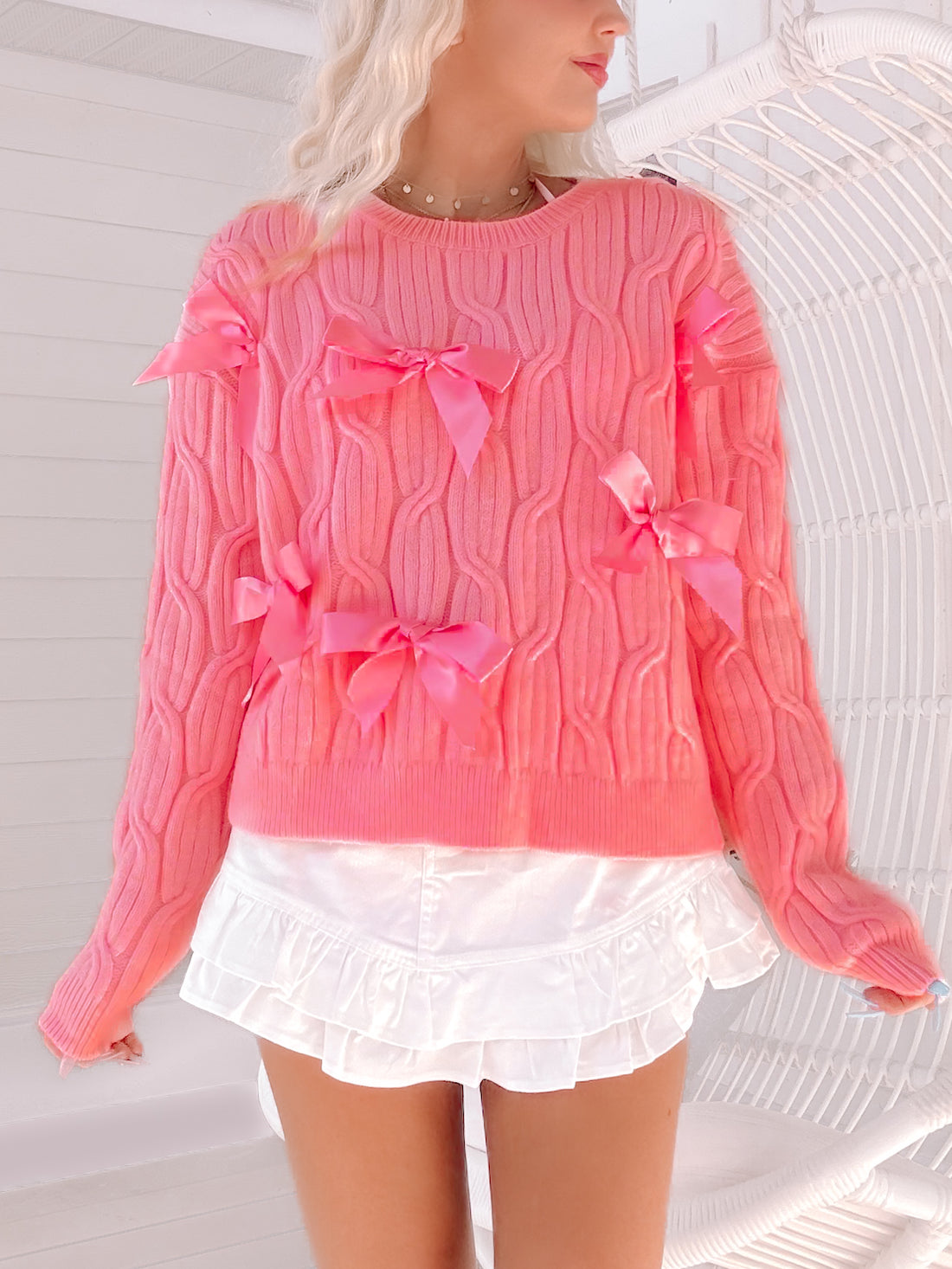 Pink cable-knit sweater adorned with pink satin bows, modeled against a white background.
