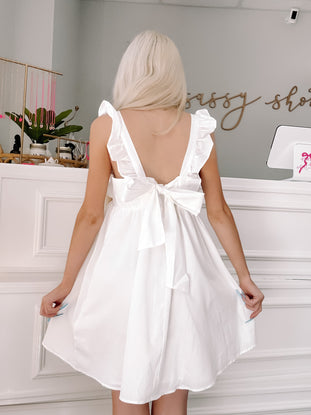 White eyelet ruffle mini dress with bow back detail, shown on model in boutique.
