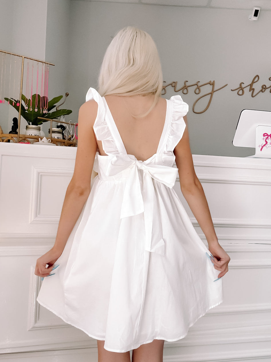 White eyelet ruffle mini dress with bow back detail, shown on model in boutique.
