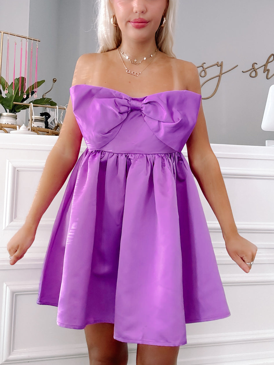 Purple satin mini dress with a large bow detail at the bodice.
