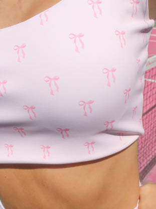 Close-up of a pale pink sports bra with a repeating pink bow print, shown on a model outdoors.
