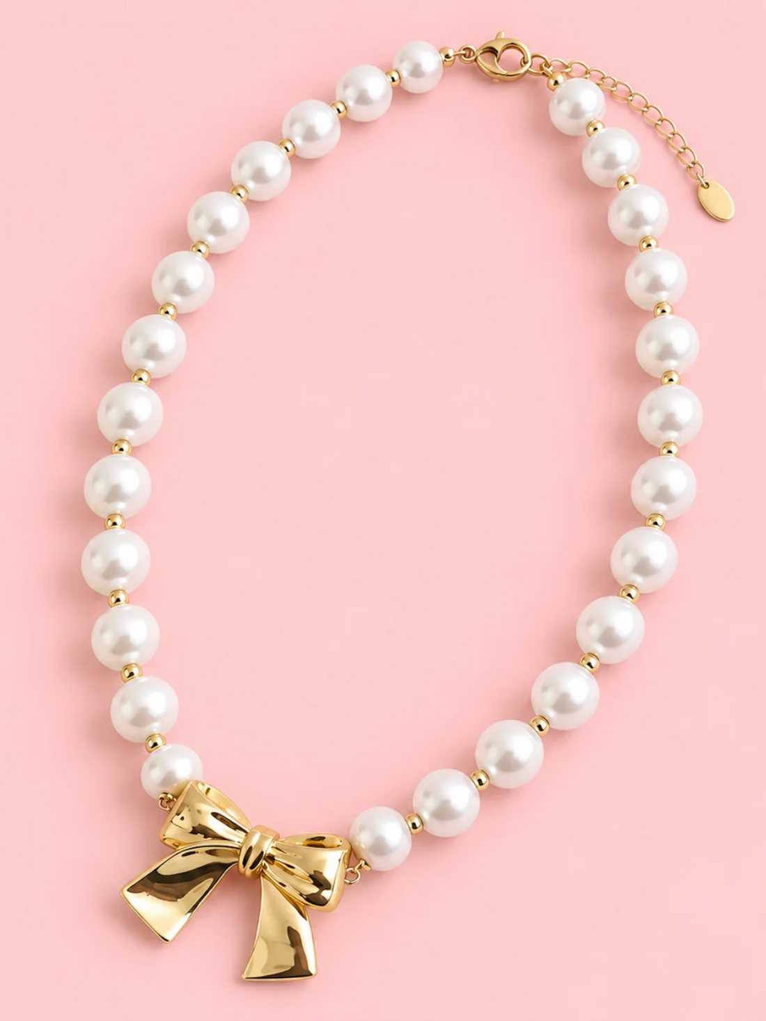 Ribbons and Pearls Necklace from Sassy Shortcake, a pearl necklace with dainty gold bead accents and a polished gold ribbon bow on a pink background.