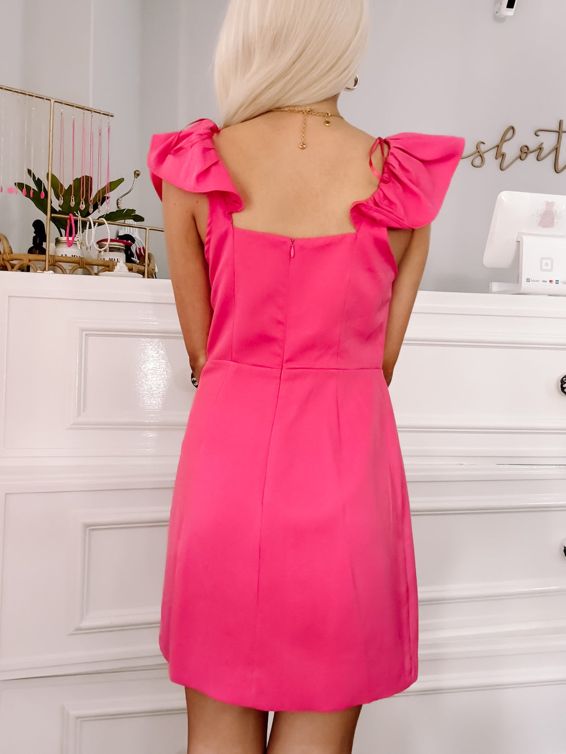Hot pink mini dress with ruffled shoulder details, shown from the back against a white dresser.
