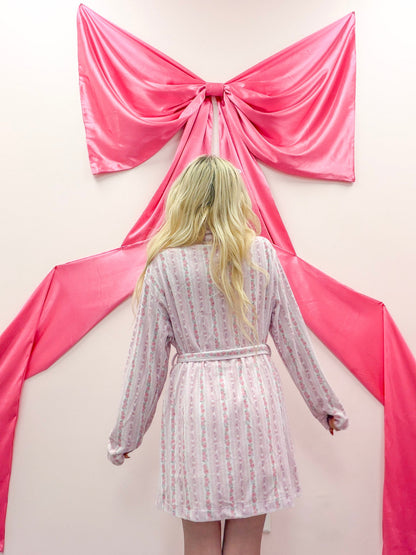 Rose Floral Robe from Sassy Shortcake - Person wearing a patterned robe standing in front of pink fabric draped like a bow on a wall.