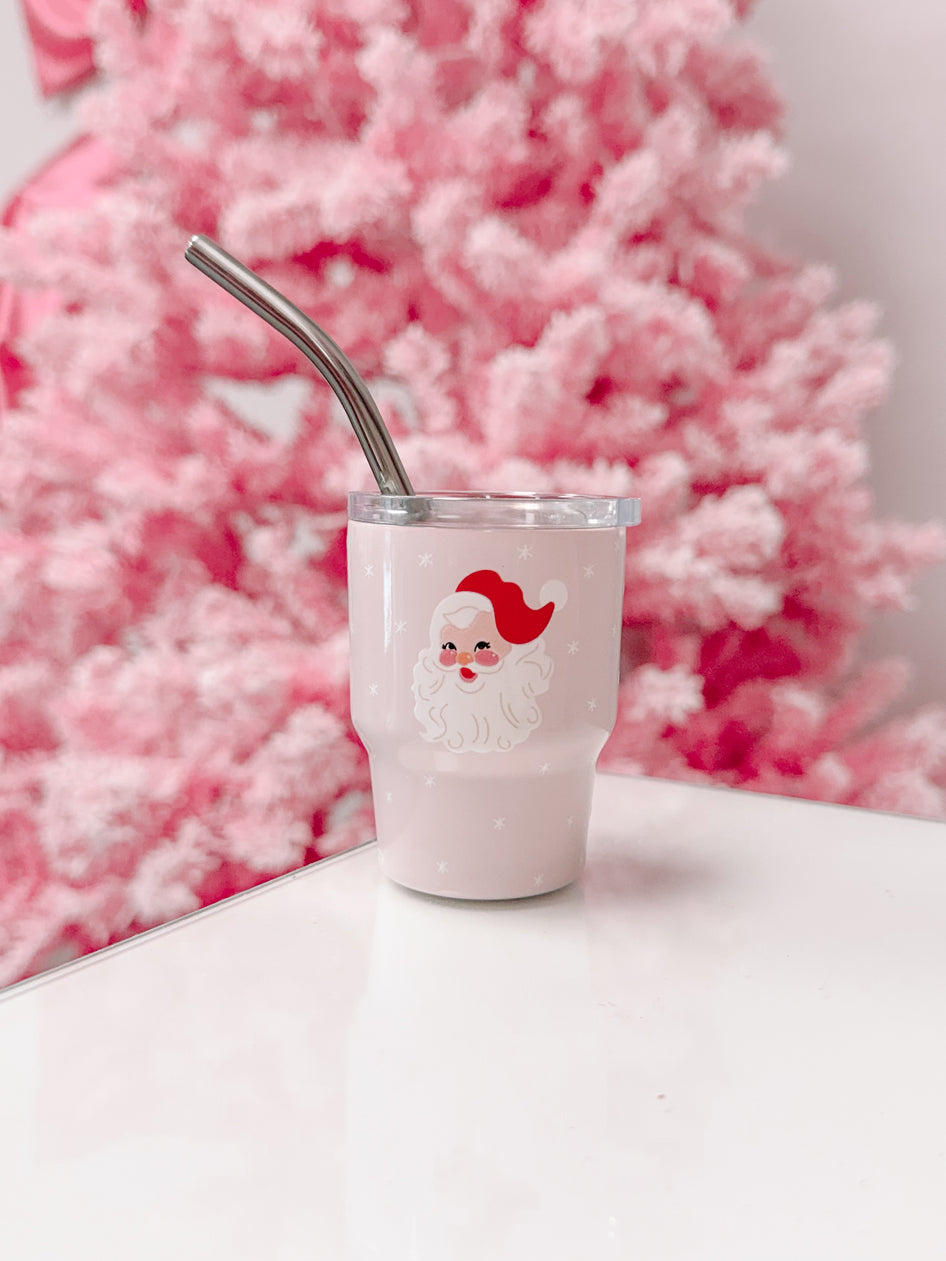 Pink Santa tumbler with metal straw, on white surface against a pink Christmas tree.
