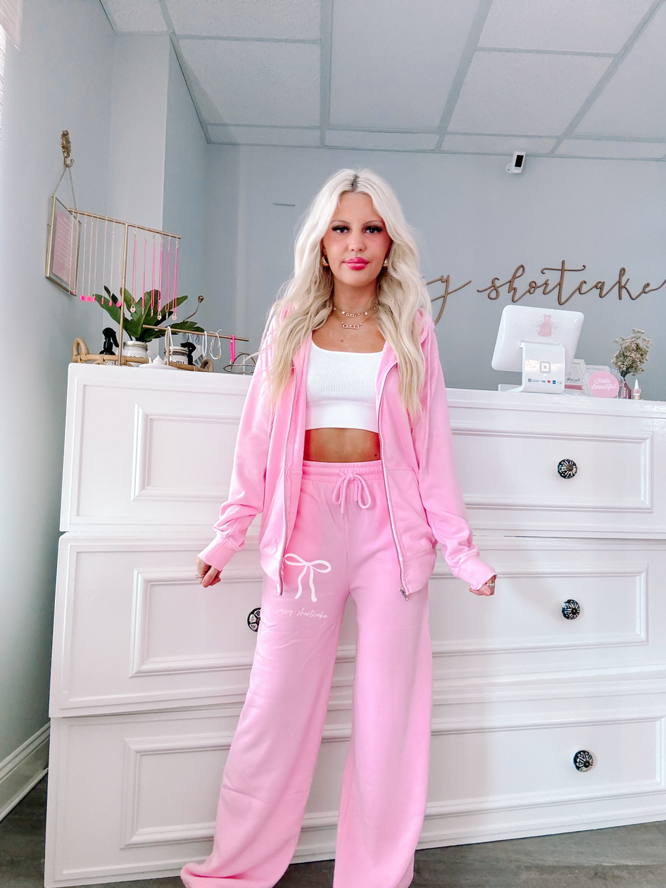Bubblegum pink wide-leg sweatpants styled with a matching zip-up hoodie, shown on a model in a boutique setting.
