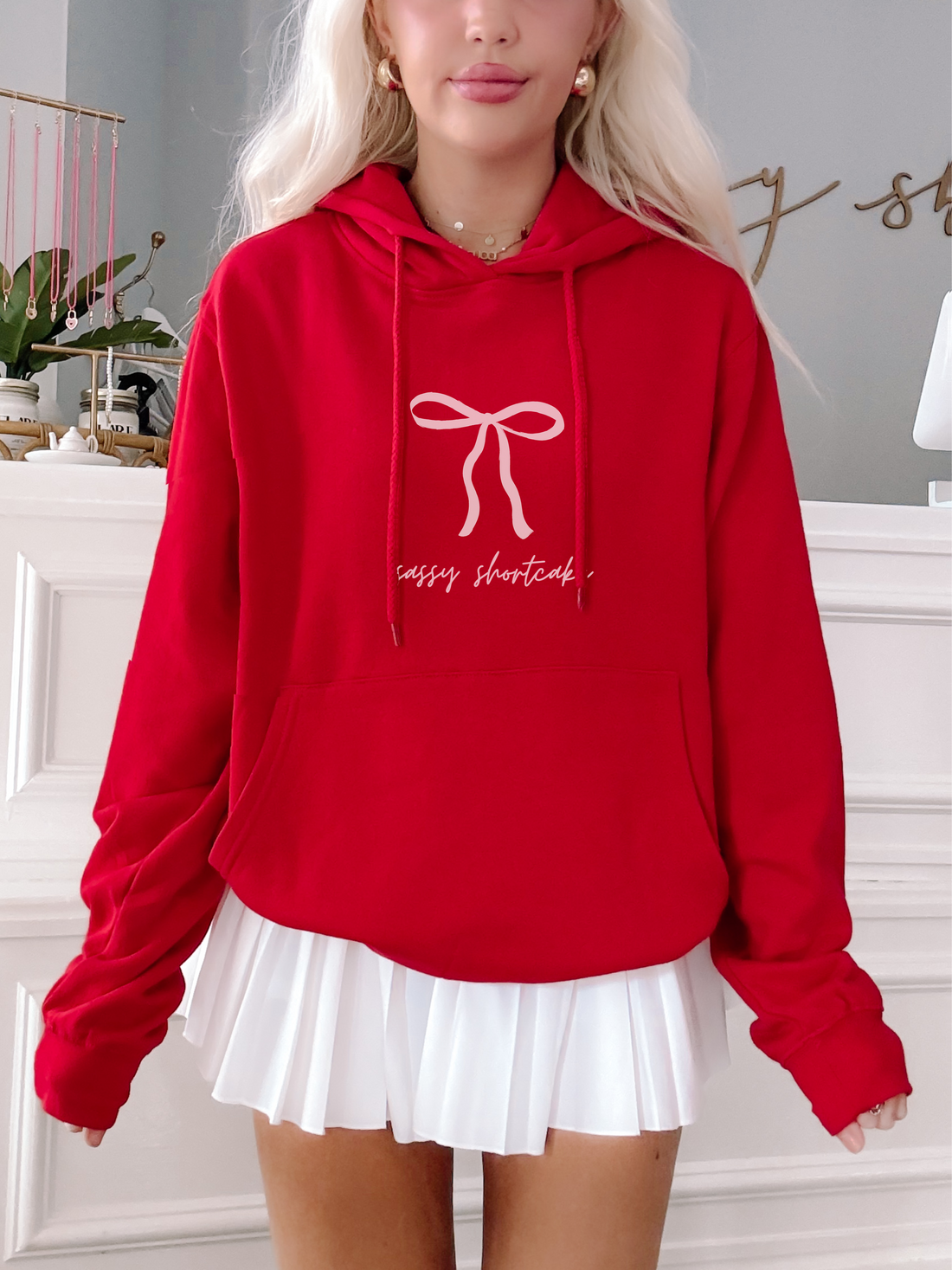 Red oversized hoodie featuring a pink bow and text graphic, paired with a white pleated skirt.
