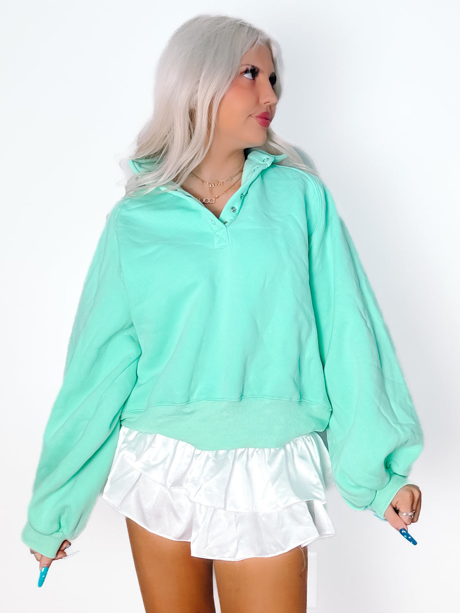 Seafoam green pullover sweater with button placket, modeled with white ruffled shorts against a white background.
