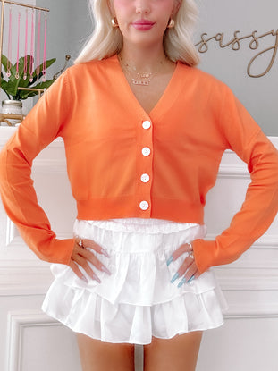 Bright orange cropped cardigan with white buttons, modeled with a white ruffled skirt.
