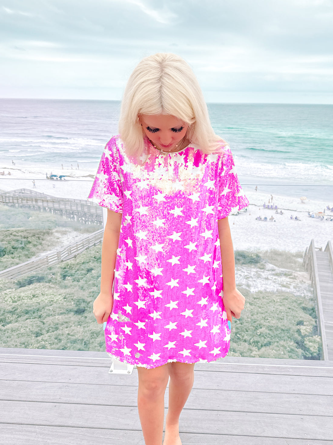 Pink sequin mini dress with white stars, shown on a model near the beach.
