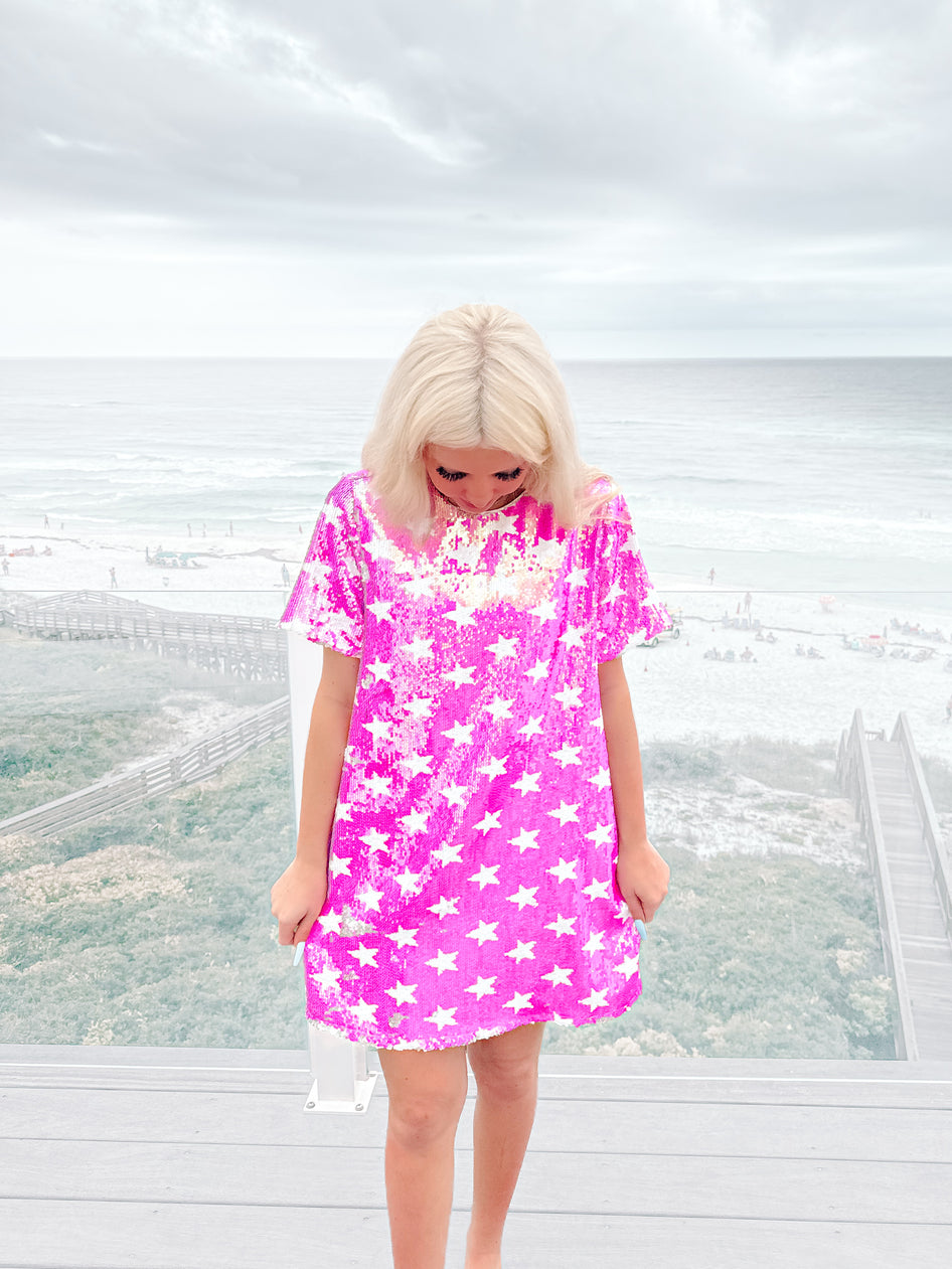 Sequined mini dress in hot pink with white stars, worn outdoors near the ocean.
