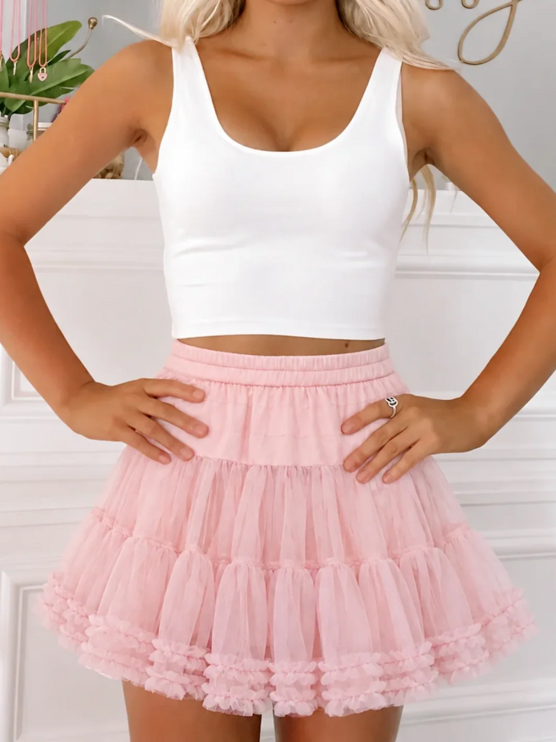 Model wearing the Sugar Plum Skirt from Sassy Shortcake, a blush pink tiered tulle mini skirt with ruffle trim and an elastic waist, styled with a white crop tank clara top.