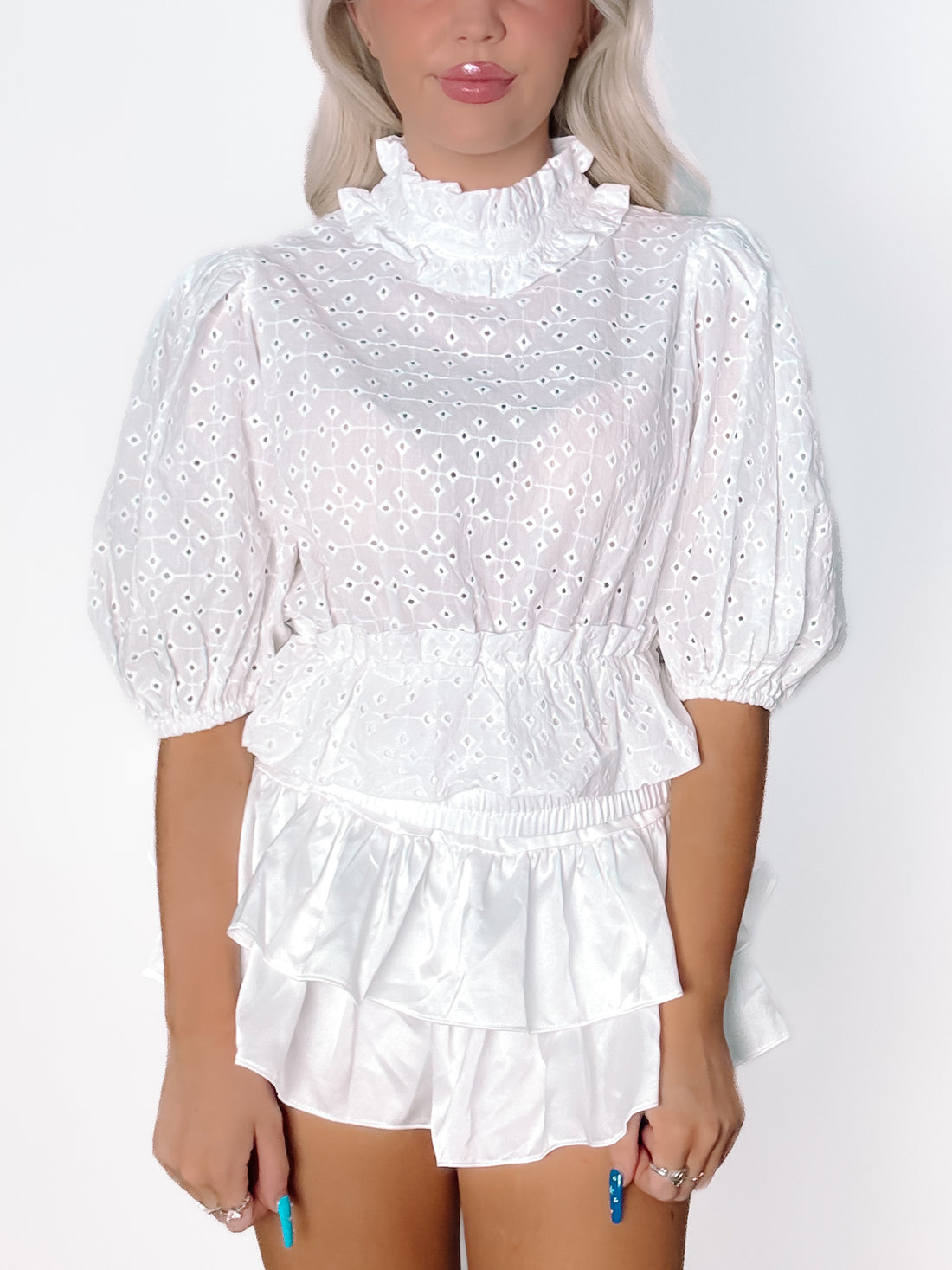 White eyelet top with puff sleeves and ruffled high neck, paired with a layered white satin mini skirt.
