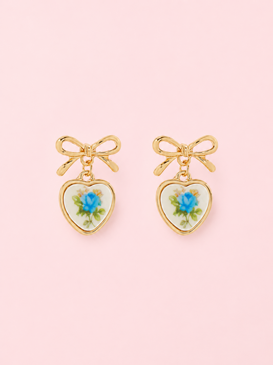 Sweet Bloom Bow Earrings in Blue from Sassy Shortcake: Gold bow earrings with ceramic heart charms featuring a blue rose print on a light pink background.