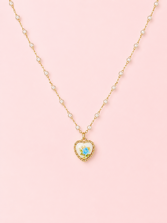 Sweet Bloom Bow Necklace in Blue from Sassy Shortcake featuring a gold-tone pearl-accent chain with a heart charm and blue rose print on a light pink background.