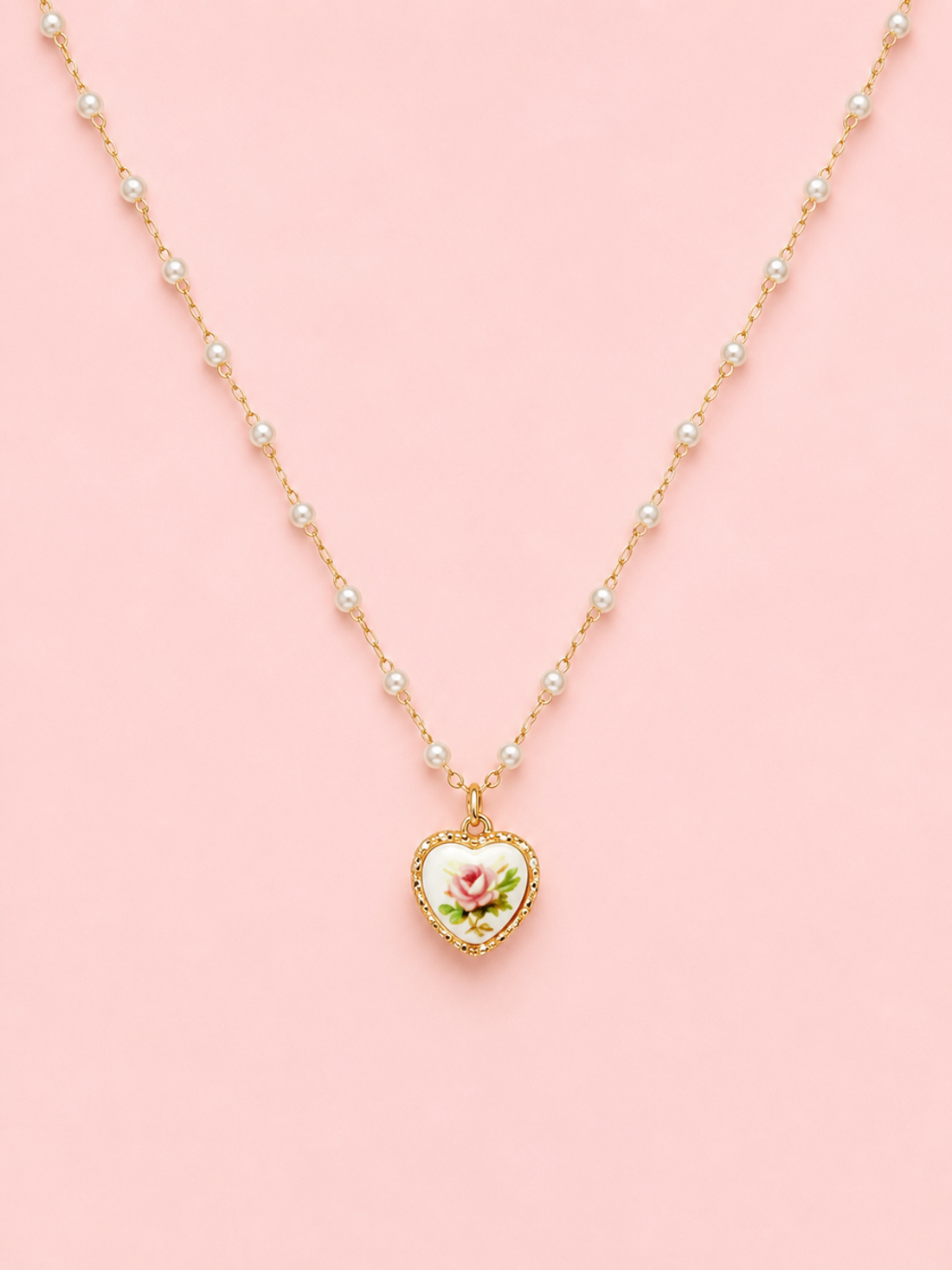Sweet Bloom Bow Necklace in Pink from Sassy Shortcake featuring a gold-tone pearl-accent chain with a heart charm and pink rose print on a light pink background.