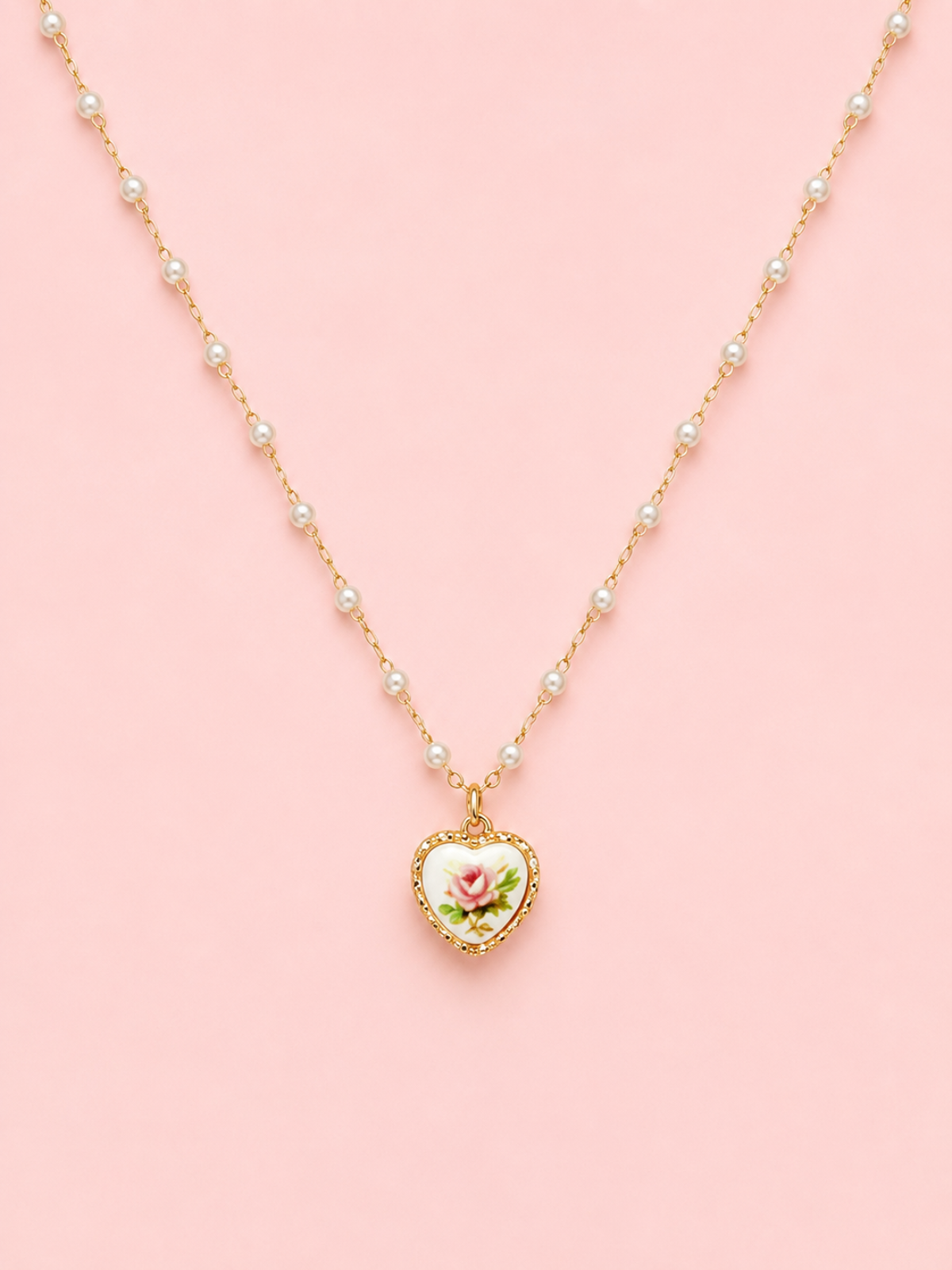 Sweet Bloom Bow Necklace in Pink from Sassy Shortcake featuring a gold-tone pearl-accent chain with a heart charm and pink rose print on a light pink background.