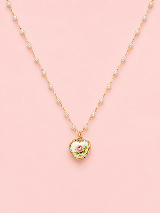 Sweet Bloom Bow Necklace in Pink from Sassy Shortcake featuring a gold-tone pearl-accent chain with a heart charm and pink rose print on a light pink background.