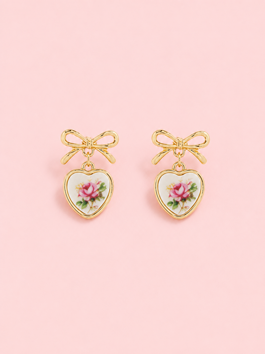 Sweet Bloom Pink Bow Earrings from Sassy Shortcake: Gold bow earrings with ceramic heart charms featuring a pink rose print on a light pink background.