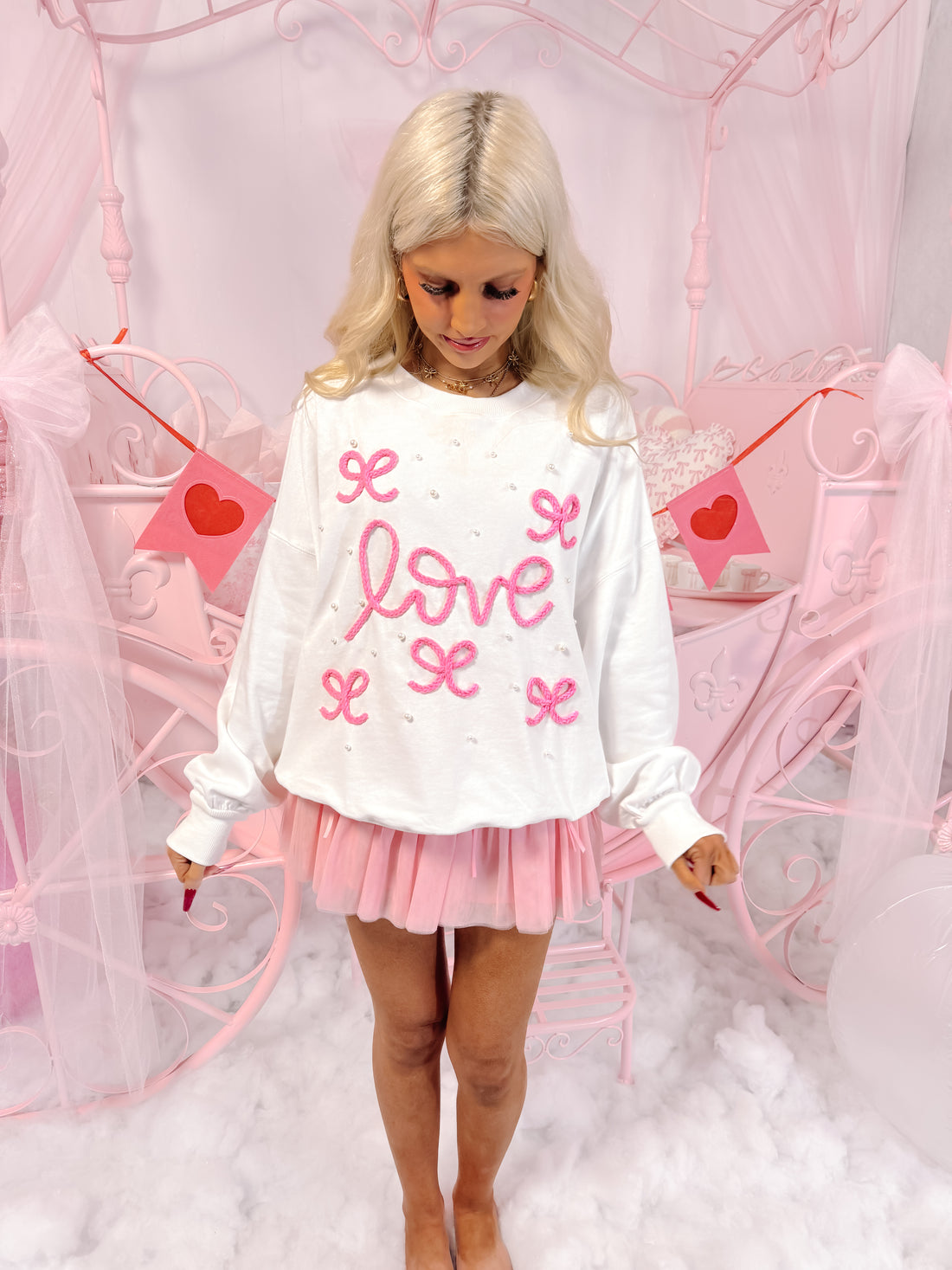 White crewneck sweatshirt with pink embroidered "love" and pearl accents, modeled against a pink backdrop.
